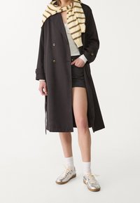Schwarzer Trenchcoat mit doppelreihigen Knöpfen, kombiniert mit einem hellgrauen Oberteil und Jeansshorts. Schal mit gelben und braunen Streifen um den Hals.