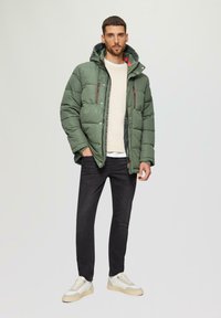 Groene pufferjack met een capuchon, voorzien van rode ritsaccenten en een gestructureerde crèmekleurige trui eronder, gecombineerd met zwarte jeans en witte sneakers.