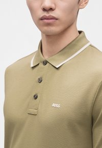 Homme portant un polo vert olive avec un col bordé de blanc et le mot "BOSS" brodé sur la poitrine.
