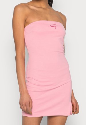 Femme portant une robe rose ajustée sans bretelles avec un petit logo rouge brodé près de la poitrine, debout devant un fond uni.