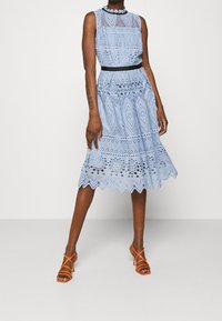 Ärmlörlös ljusblå spetsklänning med geometriska mönster och vågig nederkant, tillsammans med orangea sandaletter med klack och remmar.