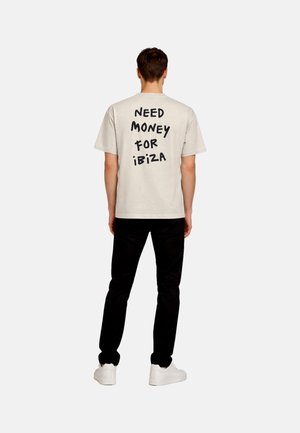 Mann steht rückwärts, trägt ein beiges Hemd mit schwarzem Text "NEED MONEY FOR IBIZA", schwarze Hose und weiße Turnschuhe vor weißem Hintergrund.