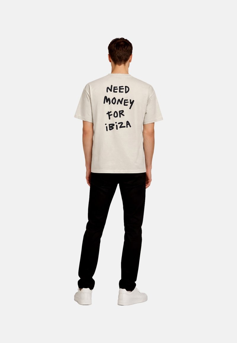 Homme debout de dos, portant une chemise beige avec un texte noir "NEED MONEY FOR IBIZA", pantalon noir et baskets blanches sur fond blanc.