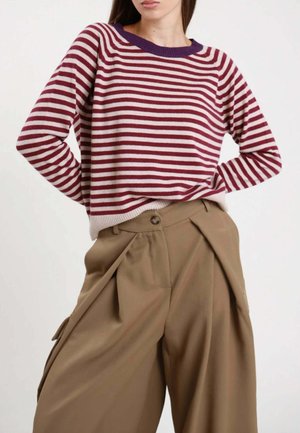 Donna che indossa un maglione a maniche lunghe a righe rosse e bianche con scollo viola e pantaloni marroni a vita alta e gamba larga.