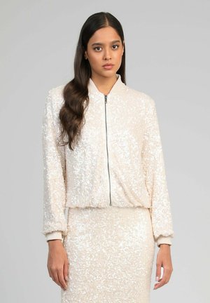 Jeune femme aux longs cheveux foncés portant une veste zippée couleur crème couverte de sequins et une jupe assortie sur un fond gris uni.