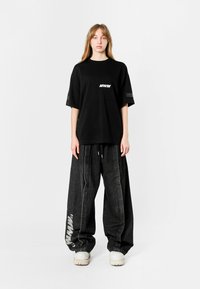 Camiseta negra de gran tamaño con el logo "MWM" en el pecho, combinada con pantalones vaqueros negros de pierna ancha con un logo impreso. Calzado blanco.