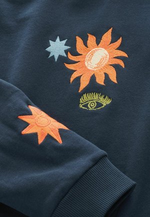 Tmavomodrá mikina s vyšívanými motívmi slnka a oka v oranžovej, žltej a svetlomodrej farbe. Mäkká textúra s rebrovanými manžetami.