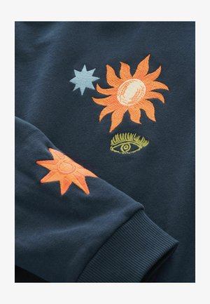 Tmavomodrá mikina s vyšívanými motívmi slnka a oka v oranžovej, žltej a svetlomodrej farbe. Mäkká textúra s rebrovanými manžetami.