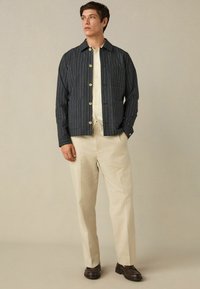 Tamno plava prugasta jakna mekog tekstura, s velikim gumbima i dva prednja džepa, u kombinaciji sa svijetlim bež hlačama i smešnim cipelama.