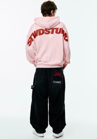 Pink hooded sweatshirt with large red "STWDSTUD" text on the back, paired with loose-fitting black denim pants featuring embroidered patches.