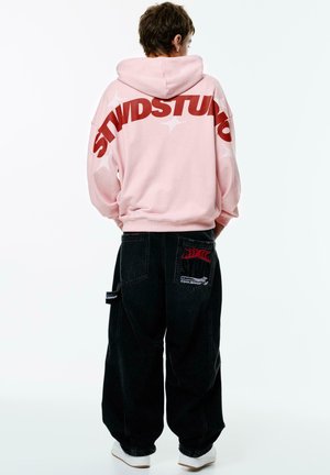 Pink hooded sweatshirt with large red "STWDSTUD" text on the back, paired with loose-fitting black denim pants featuring embroidered patches.