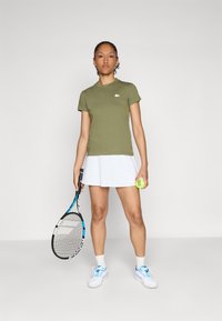 Oliven grønn bomull t-skjorte med rund hals, hvit Lacoste-logo. Kombinert med en hvit tenniskjole, holder en racket og tennisball.