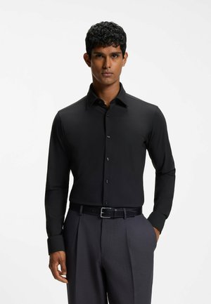 Mann steht mit den Händen in den Taschen, trägt ein eng anliegendes schwarzes langärmeliges Hemd mit Knöpfen und dunkelgraue Hose mit schwarzem Gürtel.