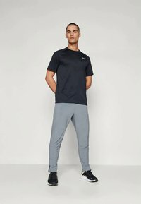 Mees seisab käed selja taga, seljas must Nike t-särk, heledad hallid spordipüksid ja mustad tossud, lihtsa tausta ees.