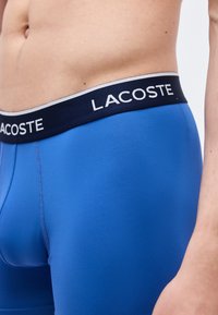 Κοντινό πλάνο ενός ατόμου που φοράει μπλε μποξεράκια Lacoste με μπλε σκούρο ελαστικό λάστιχο στη μέση που φέρει το όνομα της μάρκας σε λευκά γράμματα.