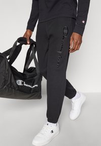 Μαύροι αθλητικοί παντελόνες με κεντητό λογότυπο "Champion" στο πλάι, σε συνδυασμό με μια μαύρη αθλητική τσάντα με λευκό λογότυπο και λευκά παπούτσια.