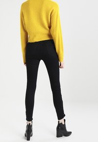 Černé skinny džíny s vysokým pasem. V kombinaci s jasně žlutým svetrem. Modelka má na sobě černé kotníkové boty na podpatku.