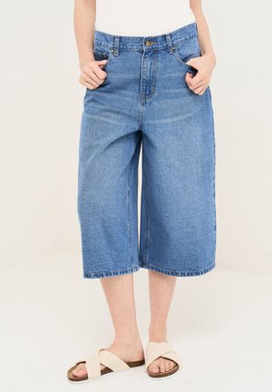 Person iført løstsiddende blå denim culottes og hvide krydssandaler, stående med hænderne i lommerne på ensfarvet baggrund.