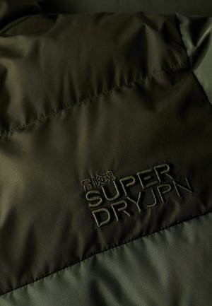 Tela acolchada verde con el logo bordado "SUPER DRYJPN". Textura suave con un brillo, que demuestra un diseño y una construcción modernos.