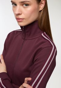 Primo piano di una donna con capelli castani lisci che indossa una giacca a zip viola con colletto alto e strisce bianche sulle maniche, con le braccia incrociate.