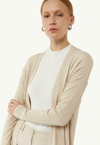 Beige Strickjacke mit gerippten Akzenten, offenes Design, leichtes Material, über einem weißen Rundhalsausschnitt getragen. Glatte Textur und weicher Abschluss.