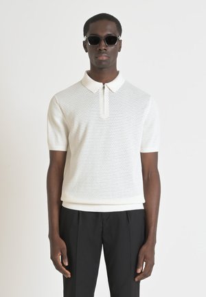 Polo shirt blanc à manches courtes avec un motif en tricot texturé et un col zippé. Association avec un pantalon noir, présente un design ajusté.