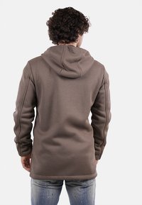 Felpa marrone a coste con maniche lunghe e cappuccio. Presenta un taglio dritto e cuciture laterali, indossata sopra un paio di jeans di denim chiaro.