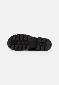 Semelle de botte noire avec des crampons profonds, comprenant des motifs en forme de flèche et un logo de marque au centre.