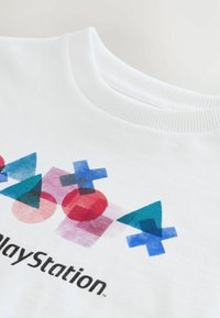 Hvid bomulds T-shirt med farverige geometriske former, herunder trekanter, cirkler, firkanter og et sort "PlayStation" logo.