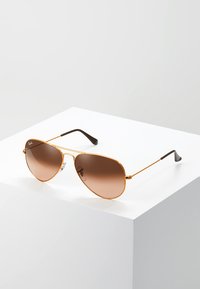 Ochelari de soare aviator cu lentile maro degradé, cadru subțire din metal aurit și vârfuri ale bradelor de culoare neagră, expuși pe un pedestal alb.