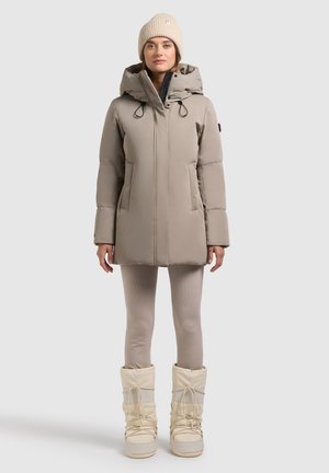 Beige geïsoleerde jas met een capuchon, voorzien van voorzakken en koordjes. Model draagt beige aangesloten leggings en crèmekleurige sneeuwlaarzen.