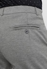 Grå byxor i texturerad tyg, med ett bakfack med knappstängning och en platt midja. Ingen synlig varumärkeslogotyp.