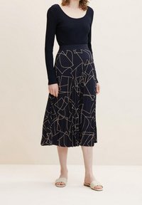 Haut côtelé à manches longues bleu marine associé à une jupe midi plissée avec un motif géométrique beige clair. Porté avec des sandales à enfiler de couleur claire.