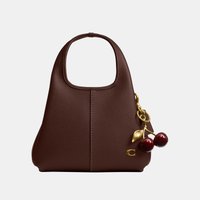 Pequeño bolso de mano marrón de cuero con asa superior y charm dorado con forma de cereza colocado cerca de la esquina inferior derecha.