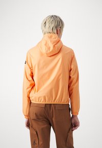 Pomarańczowa, lekka kurtka z kapturem, z wciągnięciem w talii i mankietach, zestawiona z brązowymi spodniami z bocznymi kieszeniami i logo na nogawce.