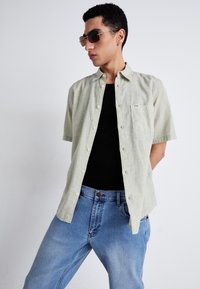 Chemise vert clair à manches courtes avec une poche sur la poitrine, assortie à un débardeur noir et un jean en denim bleu clair. Modèle portant des lunettes de soleil.