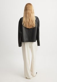 Juodas odinis švarkas su kvadratiniu dizainu, ilgomis rankovėmis ir lygiu paviršiumi, derinamas su laisvo silueto grietinėlės spalvos kelnėmis.