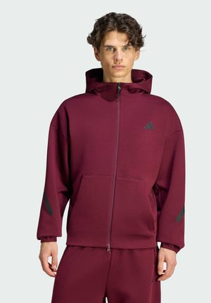 Junger Mann mit lockigem Haar, der einen weinroten Kapuzenpullover mit durchgehendem Reißverschluss und passende Hosen vor einem schlichten hellen Hintergrund trägt.