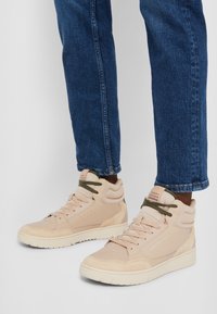Persoon in beige Tommy Hilfiger hoge sneakers met donkergroene veters, gekleed in een blauwe spijkerbroek en staande op een lichtgrijze vloer.