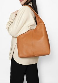 Bruine leren tote bag met een gestructureerd oppervlak, een slappe vorm en een brede schouderriem. Gefotografeerd tegen een lichte achtergrond.