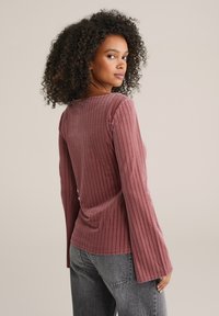 Pikkade varrukatega mauve-top, millel on tekstuurne siksakimuster ja sirge kaelus, koos kõrge vöökohaga hallide teksadega.