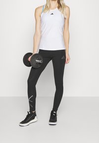 Biały sportowy top na ramiączkach z niebieskimi paskami, czarne legginsy z białymi akcentami i czarne buty sportowe, trzymający czarny hantel.