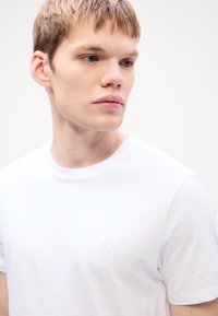 White cotton t-shirt with a crew neck and short sleeves, featuring a subtle embroidered "CK" logo on the chest. Smooth texture.