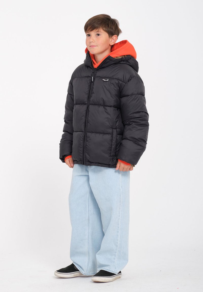 Schwarze Daunenjacke mit gesteppter Optik und Kapuze, getragen über einem orangefarbenen Hoodie. Kombiniert mit hellblauen Weitbein-Jeans und schwarzen Sneakers.