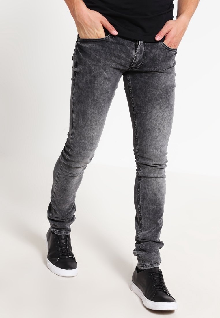 Homme portant un jean gris ajusté délavé et des baskets noires avec des semelles blanches, les mains reposant dans les poches avant.