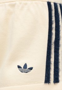 Tissu couleur crème avec logo Adidas trefoil bleu marine et trois rayures duveteuses bleu marine le long du bord.