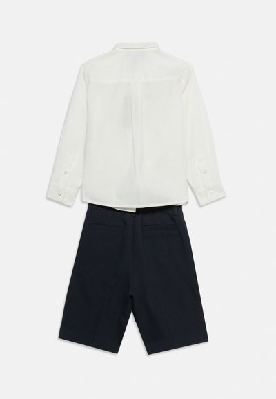 Λευκό πουκάμισο με μακριά μανίκια και κουμπιά, συνδυασμένο με μαύρο, ραμμένο σορτς μέχρι το γόνατο, απεικονισμένο από πίσω σε απλό λευκό φόντο.