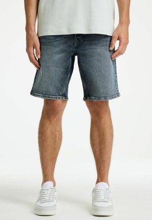 Mann trägt ausgewaschene blaue Jeansshorts, weiße Turnschuhe und ein weißes T-Shirt, steht vor einem schlichten weißen Hintergrund.