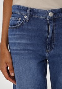 Blauwe spijkerbroek heeft een middelhoge taille, een ritssluiting aan de voorkant, een klassiek vijf-pocket ontwerp en subtiele vervaging op de stof voor textuur.
