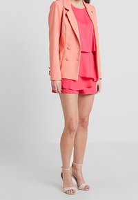 Blazer de coral sobre una blusa rosa en capas y pantalones cortos a juego; tejido suave, cierre de doble botonadura, cuello abierto, combinado con tacones beige.